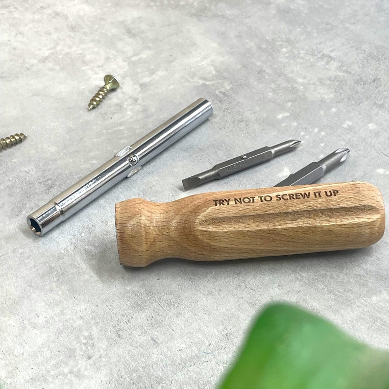 May include: A screwdriver set featuring a light brown wooden handle engraved with the text TRY NOT TO SCREW IT UP, a silver metal shaft, and two interchangeable double-ended screwdriver bits, accompanied by two small metal screws.
