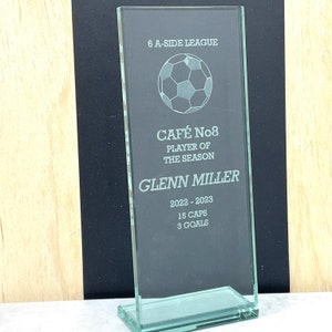 May include: A green glass trophy with a soccer ball design. The text on the trophy reads "6 A-SIDE LEAGUE", "CAFÉ No8", "PLAYER OF THE SEASON", "GLENN MILLER", "2022 - 2023", "15 CAPS", and "3 GOALS".