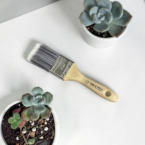 May include: A paint brush with a light wood handle and a metal ferrule, featuring synthetic bristles. The handle is printed with a house icon and the text "TOM & KELLY HAPPY NEW HOME". The metal ferrule is clearly marked with "50mm 2in".