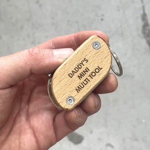 Pode incluir: Um chaveiro de madeira com um anel de prata. O chaveiro é gravado com o texto "Daddy's Mini Multi Tool".