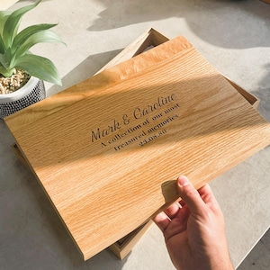 Puede incluir: Una caja de madera clara con tapa, con texto grabado que dice "Mark & Caroline, A collection of our most treasured memories, 23.08.86." La caja está abierta, revelando el interior. Una mano sostiene la tapa.