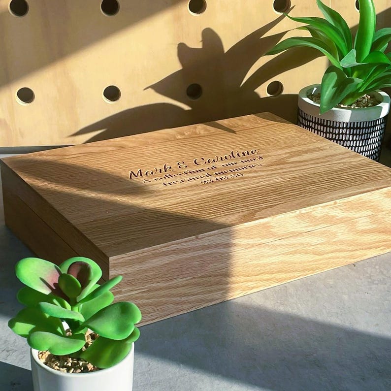 Pode incluir: Uma caixa de madeira castanha clara, de forma retangular, com uma tampa gravada com o texto "Mark & Caroline". Pequenas plantas em vasos rodeiam a caixa.
