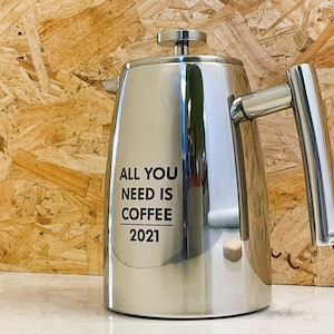 May include: A shiny stainless steel coffee press with a long handle and a lid with a small knob. The words "ALL YOU NEED IS COFFEE 2021" are printed in black on the front. The coffee press is designed for brewing coffee.