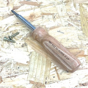 May include: A screwdriver with a polished metal shaft and a light brown wooden handle featuring the engraved text TRY NOT TO SCREW IT UP. Two small silver metal screws are positioned to the left of the tool on the surface.