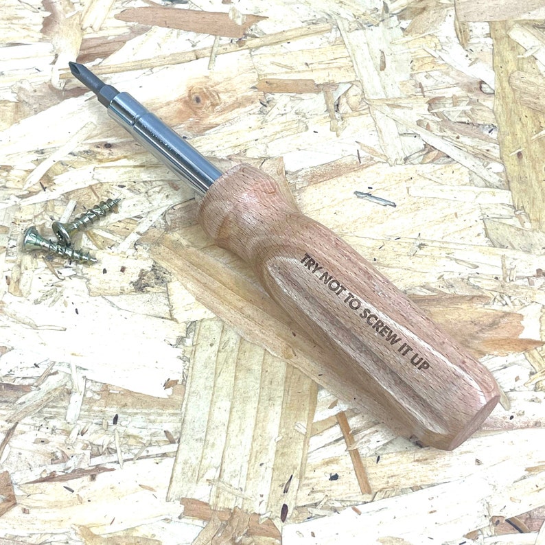 May include: A screwdriver with a polished metal shaft and a light brown wooden handle featuring the engraved text TRY NOT TO SCREW IT UP. Two small silver metal screws are positioned to the left of the tool on the surface.
