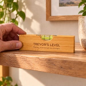 May include: A rectangular bamboo spirit level featuring a central green liquid vial. The wood surface is engraved with the text "TREVOR'S LEVEL" and "Making sure things are straight" in a dark brown sans-serif font.