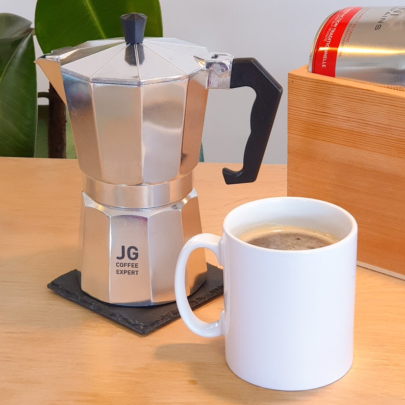 May include: A silver stovetop espresso maker with a black handle and lid sits on a square coaster. A white mug filled with coffee is next to the espresso maker. The text "JG COFFEE EXPERT" is printed on the espresso maker.