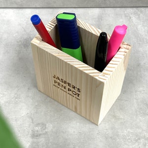 Könnte beinhalten: Ein hölzerner Stiftehalter mit dem Text "Jasper's Pen Pot Keep Your Desk Tidy" auf der Vorderseite. Der Halter ist mit einem rosa Textmarker, einem blauen Stift und einem schwarzen Stift gefüllt.