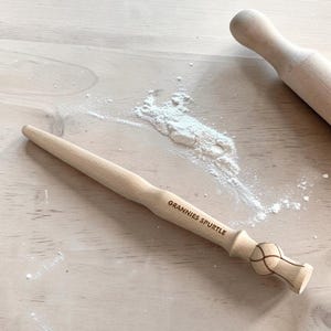 Può includere: Uno spurtle in legno chiaro con manico affusolato e base arrotondata caratterizzata da un motivo a nodo inciso. Il testo "GRANNIES SPURTLE" è impresso sul corpo. Sono visibili una parte di un mattarello in legno e della farina bianca.