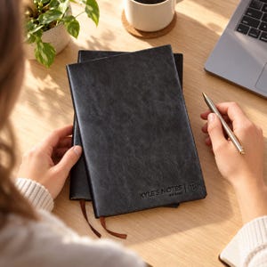 May include: A black notebook with a textured cover featuring the embossed text KYLE'S NOTES THE BOSS and a small circular logo. The notebook has a brown ribbon bookmark extending from the bottom edge and is held alongside a silver metal pen.