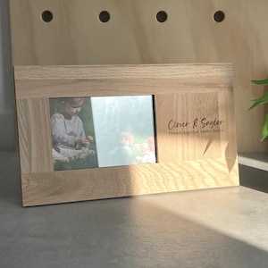 May include: A wooden photo frame with a light brown finish. The frame has a cutout for a photo and a personalised inscription that reads "Clover & Saylor, Friends together, sisters forever".