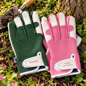 May include: A pair of gardening gloves, one dark green and one pink, featuring white leather-like accents on the fingers and palms. The green glove has the word Grandad printed on the wrist strap, while the pink glove has the text To Mum love Olivia x.