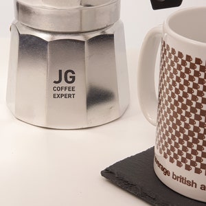 May include: A silver stovetop espresso maker with the text "JG COFFEE EXPERT" is next to a white mug with a brown checkered pattern. A black slate coaster is in the foreground.