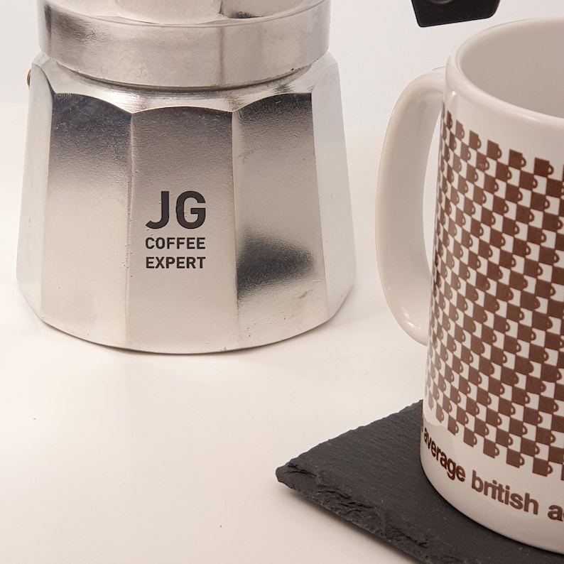 May include: A silver stovetop espresso maker with the text "JG COFFEE EXPERT" is next to a white mug with a brown checkered pattern. A black slate coaster is in the foreground.