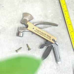 Könnte beinhalten: Ein Multitool mit einem Holzgriff, der mit "Tom's Multi Tool For Wild Camping and Odd Jobs" graviert ist. Das Werkzeug verfügt über einen Hammer, eine Axt, ein Messer und andere Werkzeuge.