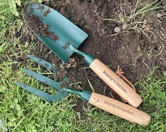 May include: A dark green metal hand trowel and a matching three-tine hand cultivator, both featuring light-colored wooden handles engraved with the text Mandy's Garden and attached brown leather hanging loops for storage.