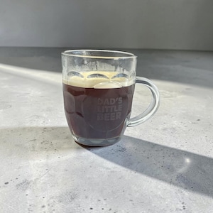 May include: A clear glass beer mug with a sturdy handle and a dimpled exterior design. The glass features the etched text DAD'S LITTLE BEER on the front. The mug is filled with a dark brown liquid topped with a thin layer of light-colored foam.