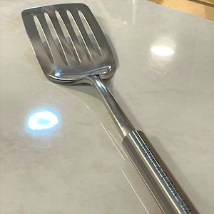 May include: A stainless steel slotted spatula with a long handle. The spatula head has multiple slots and a curved design. The handle has the text "YOU'RE A FLIPPIN' STAR" engraved on it.