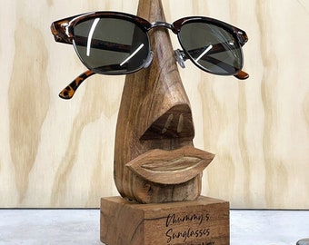 Puede incluir: Portagafas de madera con forma de cara con nariz, labios y ojos. El soporte tiene un acabado de madera marrón y está grabado con el texto "Thummy's Sunglasses Lots of love from Clover & Spice".