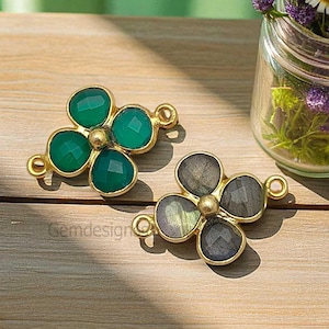 May include: Two flower-shaped gemstone pendants. One features green gemstones, the other gray. Both have gold-colored metal settings and small loops for attaching to jewelry. The pendants are displayed on a wooden surface.