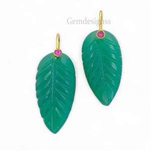 Puede incluir: Un par de pendientes verdes con forma de hoja con detalles tallados. Cada pendiente presenta una pequeña gema rosa redonda y un gancho dorado. Los pendientes están sobre un fondo blanco. El texto "Gemdesignss" es visible.