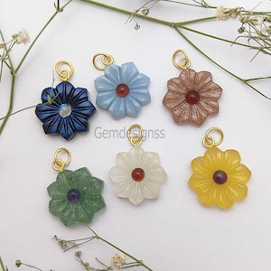 Könnte beinhalten: Sechs blütenförmige Edelsteinanhänger in verschiedenen Farben: Marineblau, Hellblau, Braun, Grün, Weiß und Gelb. Jeder Anhänger hat eine goldfarbene Schlaufe und einen zentralen Edelstein. Die Anhänger werden auf einer weißen Oberfläche mit Zweigen weißer Blumen präsentiert.