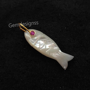 Może przedstawiać: Biała zawieszka w kształcie ryby z wytrawionymi detalami i złotą pętlą. Ryba ma czerwony klejnot w oku. Zawieszka jest na czarnym tle. Tekst "Gemdesignss" znajduje się w lewym górnym rogu.