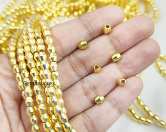 Pode incluir: Um fio de contas de metal dourado com formato cilíndrico facetado. As contas são enfiadas em um fio e têm um diâmetro aproximado de 6 mm. Há também várias contas soltas na imagem.