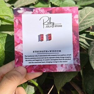 Puede incluir: Un par de pendientes de rubí con engastes de oro. Los pendientes están en una tarjeta blanca con las palabras "Ruby July Stone" y "Strength + Wisdom". La tarjeta también incluye una descripción de la piedra de rubí.