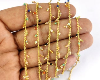 May include: Gold chain with colorful gemstone beads hanging from it. The chain is on a hand, and the beads are various colors, including red, green, blue, and pink.
