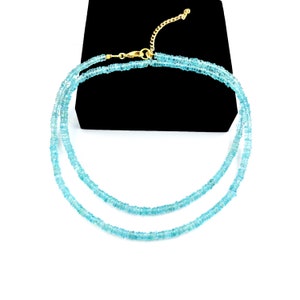 May include: A delicate, double-strand necklace featuring small, light blue beads. The necklace has a gold-colored clasp and chain. The jewelry is displayed against a black background and a white surface.