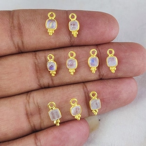 May include: Gold-coloured rectangular charms with a small loop at the top and a cluster of three small beads at the bottom. Each charm has a square, iridescent, light-coloured stone set in the centre. The charms are displayed on a light-coloured surface.