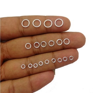 May include: A collection of small, silver-colored jump rings of varying sizes, arranged on a person's fingers. The rings are round and appear to be made of metal. The image is well-lit, showing the details of the rings.