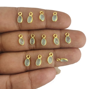 Op de afbeelding: Een verzameling van 12 vergulde ovale bedels met een lichtblauwe edelsteen. Elke bedel heeft een lus om aan sieraden te bevestigen.