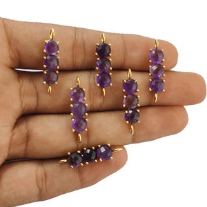 Può includere: Sei ciondoli dorati con tre pietre rotonde di ametista ciascuno. I ciondoli sono disposti su una mano.