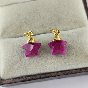 Pode incluir: Dois pingentes de estrela rosa com forma de estrela com fio dourado enrolado ao redor da parte superior. Os pingentes são exibidos em uma caixa de joias forrada de veludo.