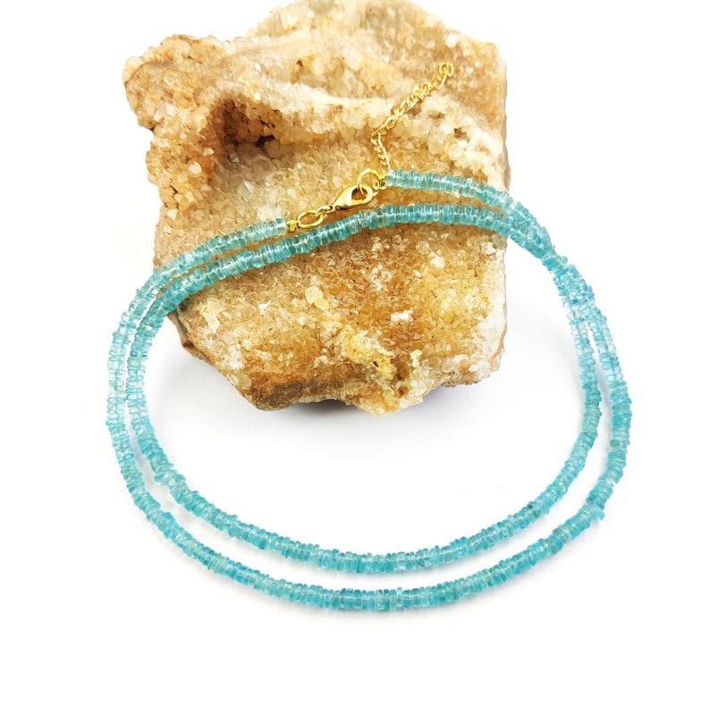 May include: A delicate necklace featuring small, light blue beads strung together. The necklace has a gold-colored clasp and chain. The beads are evenly spaced, creating a smooth, elegant look. The necklace is displayed on a textured, tan-colored surface.