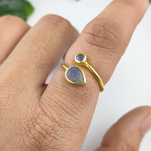 Pode incluir: Anel de ouro com duas pedras de labradorita em forma de lágrima. A pedra maior tem a forma de uma lágrima e a menor é redonda. O anel é ajustável.