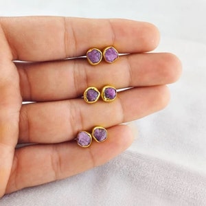Puede incluir: Un par de pendientes de oro con peque&ntilde;as piedras preciosas p&uacute;rpuras irregulares. Los pendientes se muestran en una mano.