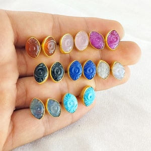 Puede incluir: Un conjunto de 12 pares de pendientes de tono dorado con piedras preciosas ovaladas que presentan un diseño de ojo. Las piedras preciosas son de varios colores, incluidos el negro, el azul, el rosa, el rojo y el blanco.
