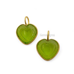May include: Two heart-shaped pendants. Each has a light green gemstone centre and a gold-coloured metal frame. A small loop at the top of each pendant is for hanging. The gemstone has a heart design carved into the centre. The text "Gemdesignss" is visible.