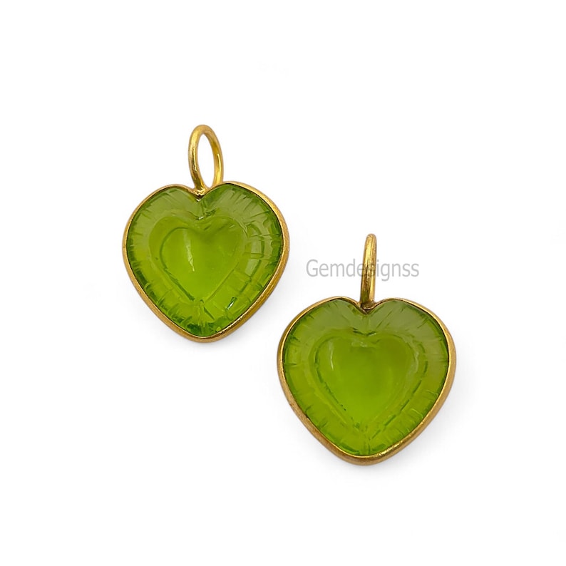 May include: Two heart-shaped pendants. Each has a light green gemstone centre and a gold-coloured metal frame. A small loop at the top of each pendant is for hanging. The gemstone has a heart design carved into the centre. The text "Gemdesignss" is visible.