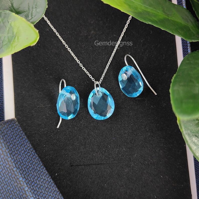 May include: A silver necklace and earrings set with light blue gemstone pendants. The gemstones are faceted and have a teardrop shape.