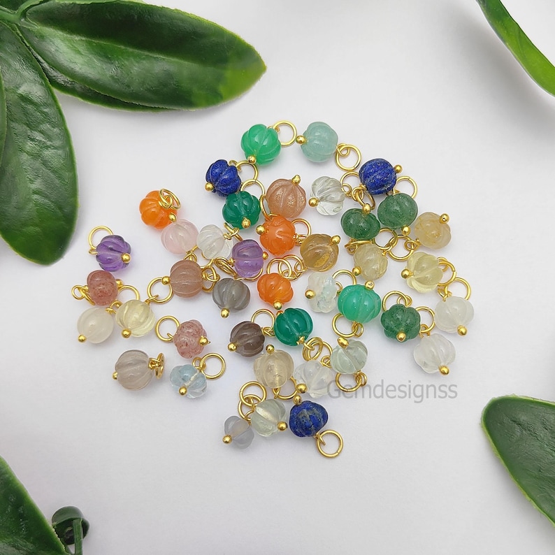 May include: A collection of colorful gemstone beads in various shades of green, orange, purple, and clear, with gold-colored metal loops. The beads are pumpkin-shaped and arranged on a white surface with green leaves.