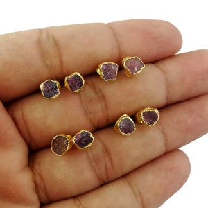 May include: Six gold-plated stud earrings, each featuring a rough-cut, dark pink gemstone. The earrings are displayed in two rows of three. The gold settings highlight the natural, irregular shape of the pink stones. A simple, elegant jewellery design.