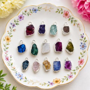Puede incluir: Una colección de catorce colgantes de piedras preciosas en bruto e irregulares con engarces de metal plateado. Cada colgante presenta una piedra única de corte irregular en varios colores, incluyendo rojo oscuro, morado, azul claro, marrón, azul oscuro, verde, blanco y amarillo.