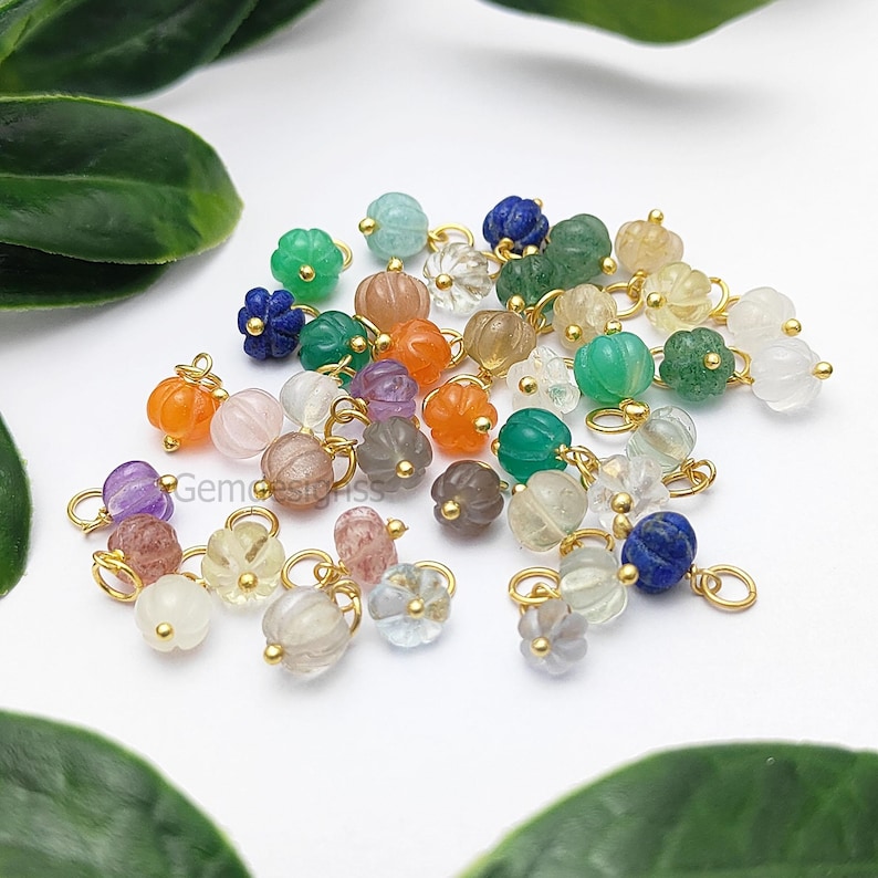 May include: A collection of colorful gemstone beads in various shapes and sizes, including pumpkin and flower designs. The beads are in shades of orange, green, purple, and clear, with gold-colored metal loops for stringing. The beads are scattered on a white surface.