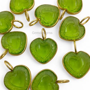 May include: A collection of heart-shaped pendants in a vivid green colour. Each pendant has a heart design etched within and is encased in a gold-coloured metal frame. They have a loop at the top for suspension. The brand name "Gemdesignss" is visible.