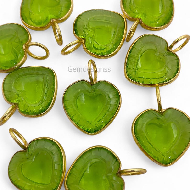 May include: A collection of heart-shaped pendants in a vivid green colour. Each pendant has a heart design etched within and is encased in a gold-coloured metal frame. They have a loop at the top for suspension. The brand name "Gemdesignss" is visible.