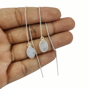 Puede incluir: Un par de pendientes de cadena de plata con un colgante de piedra ovalada de color púrpura claro en cada cadena. Los pendientes se muestran en una mano.